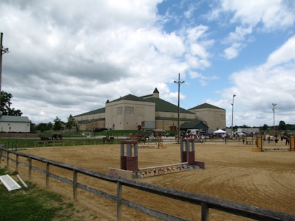 A large, beige equestrian building with green-roofed architecture is surrounded by an outdoor horse arena. Horses and riders are visible in the ring. The sky is partially cloudy, and the setting appears to be a horse show or event.