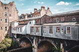 An architectural scene featuring historic stone buildings with multiple chimneys and tall windows, constructed on an old bridge. The structure spans over a river or canal, with lush greenery visible below. The weathered appearance and rustic colors convey a sense of history.