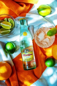 A colorful composition featuring a bottle of mineral water on a vibrant orange and white towel, surrounded by whole and sliced citrus fruits such as limes and oranges. A glass with ice and a slice of lime is also present.