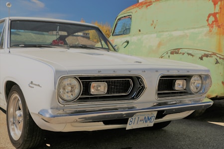 A vintage white Plymouth Barracuda car is parked next to an old, weathered green truck with patches of rust. The image captures the front side of the Plymouth, showcasing its classic dual headlight design and chrome details. The scene suggests a sunny day with a clear blue sky in the background.