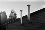 Two metal chimneys extend from the roof of a wooden structure against a backdrop of a clear sky and trees. The scene is depicted in black and white, creating strong contrasts between the building, trees, and the sky.