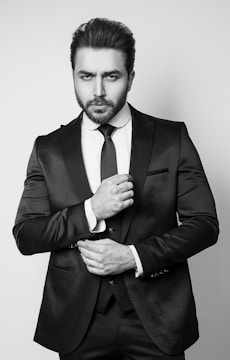 A man stands in a formal pose wearing a dark, textured suit with a crisp white shirt and a tie. His facial expression is serious, and he adjusts the front of his suit jacket with one hand.