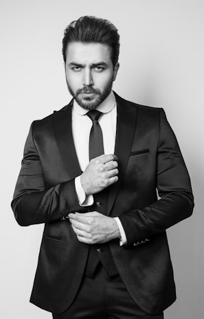 A man stands in a formal pose wearing a dark, textured suit with a crisp white shirt and a tie. His facial expression is serious, and he adjusts the front of his suit jacket with one hand.