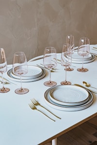 A neatly arranged dining table with elegantly placed dinnerware and glasses. The table setting includes plates stacked on top of each other, accompanied by forks, spoons, and knives, all made of gold or brass material. Wine glasses with long stems are symmetrically aligned, creating a sophisticated and formal dining atmosphere. The table has a light-colored surface, and the background wall has a textured, neutral tone.