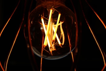 A close-up view of a glowing incandescent light bulb with visible filaments casting warm, orange light against a dark background. The intricate design and reflections on the bulb surface enhance the luminous effect.