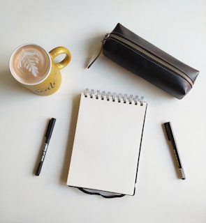 white ceramic mug with coffee beside black pen and white notebook