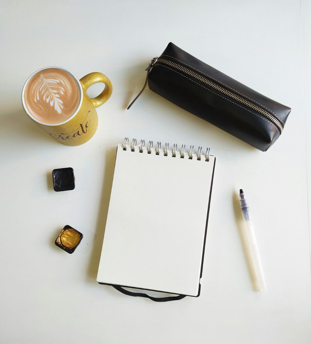 Tasse en céramique blanche à côté d’une pochette en cuir noir et d’un stylo à clic blanc