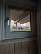 A window view shows a slanted roof overhang and the exterior of a house with a dark roof and white trim. The sky is overcast with orange and blue hues, and there are tall grasses visible near the house.