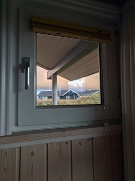 A window view shows a slanted roof overhang and the exterior of a house with a dark roof and white trim. The sky is overcast with orange and blue hues, and there are tall grasses visible near the house.