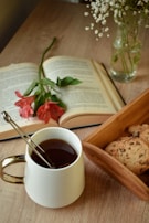A cozy arrangement featuring an open book with a red flower resting on it, a white mug filled with a dark beverage, and a small wooden tray containing chocolate chip cookies. In the background, a vase holds delicate white flowers, all set on a wooden table.
