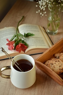 A cozy arrangement featuring an open book with a red flower resting on it, a white mug filled with a dark beverage, and a small wooden tray containing chocolate chip cookies. In the background, a vase holds delicate white flowers, all set on a wooden table.