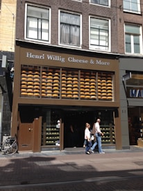 A storefront labeled 'Henri Willig Cheese & More' with large displays of stacked cheese wheels behind glass. The building has several windows, and there are people walking by on the sidewalk. A bicycle is parked on the left side.