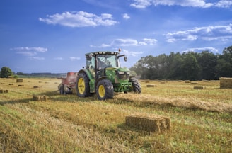 A green tractor is driving through a field, pulling a red hay baler that is creating rectangular bales of hay. The field is filled with scattered hay bales, and a line of trees can be seen in the background under a blue sky with scattered clouds.