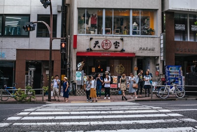 A group of people stands on the sidewalk of an urban street, watching the traffic light above a pedestrian crossing. They are in front of a vibrant street with signs in different languages, indicating a cultural or commercial area. The street features a restaurant with a recognizable red and white sign, and there are bicycles parked nearby and vending machines visible.