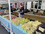 A bustling farmers market stand features a variety of fresh vegetables displayed in an orderly arrangement. Basketfuls of potatoes, different types of beans, and other produce are laid out across colorful, patterned tablecloths. A vendor wearing a cap, gloves, and a mask stands behind the stall, assisting customers.