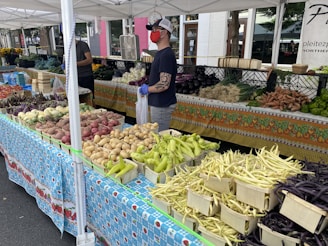 A bustling farmers market stand features a variety of fresh vegetables displayed in an orderly arrangement. Basketfuls of potatoes, different types of beans, and other produce are laid out across colorful, patterned tablecloths. A vendor wearing a cap, gloves, and a mask stands behind the stall, assisting customers.