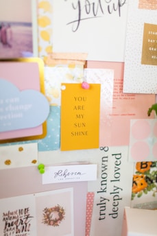 A collage of various inspirational and decorative pieces, featuring a central yellow card that reads 'YOU ARE MY SUNSHINE' surrounded by other cards with positive quotes and pastel colors. The arrangement includes elements like floral patterns, calligraphy, and soft textures, pinned to a board.