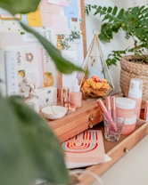 A cozy, aesthetically arranged vanity table with a selection of skincare products, makeup items, and decorative objects. The table features wooden drawers and is adorned with a potted plant. On the board behind are various pinned notes and cards, adding a creative, personal touch. The display includes a geometric jewelry holder and vibrant decorative elements.