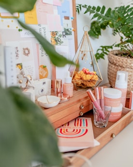 A cozy, aesthetically arranged vanity table with a selection of skincare products, makeup items, and decorative objects. The table features wooden drawers and is adorned with a potted plant. On the board behind are various pinned notes and cards, adding a creative, personal touch. The display includes a geometric jewelry holder and vibrant decorative elements.