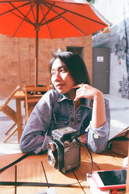 A warm, candid photo of a sunlit outdoor workspace with a camera and notebook resting on a wooden table.