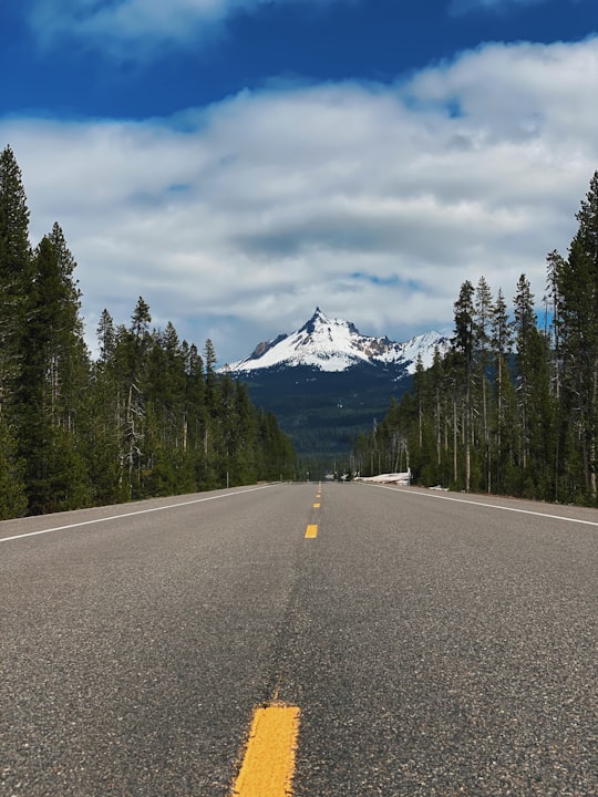 gray asphalt road between green trees under white clouds and blue sky during daytime in Mount Thielsen United States