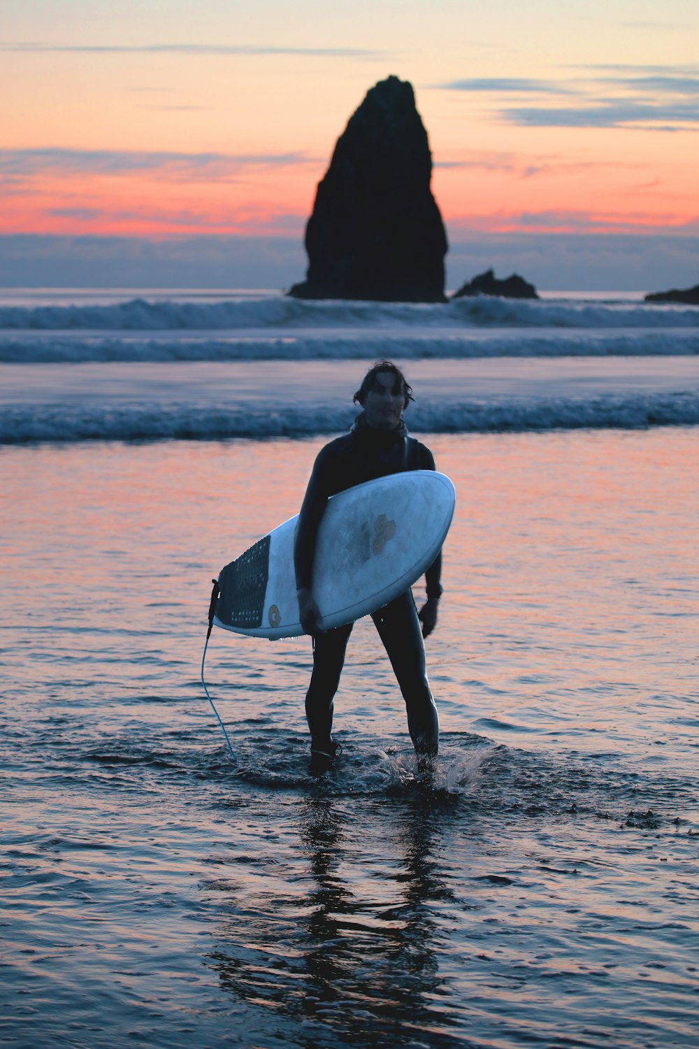 femme en combinaison de plongée noire tenant une planche de surf blanche marchant sur le bord de mer pendant la journée