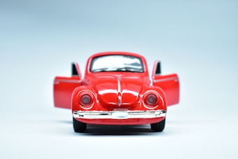 A toy model of a classic red car with its doors open, positioned against a smooth, light blue background. The car is viewed from the front, highlighting its vintage design elements such as rounded headlights and a shiny chrome bumper.