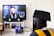 A student wearing a black graduation cap and gown watches a virtual commencement ceremony on a television screen. The scene is set in a living room with books on a white shelf underneath the TV and a laptop also displaying the ceremony.