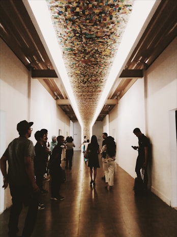 A long, white-walled corridor featuring an artistic, colorful ceiling installation. Several people are walking or standing along the hallway, engaged in casual conversation or looking at their phones. The floor is made of wood, adding warmth to the otherwise minimalist space.
