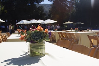 A beautifully arranged outdoor event featuring long tables adorned with floral centerpieces. The tables, covered in white tablecloths, are lined with wooden chairs. In the background, groups of people gather under large white umbrellas in a sunlit setting surrounded by trees.
