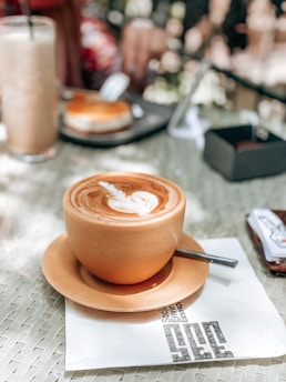 A beautifully crafted cup of coffee with latte art sits on a saucer atop a napkin on a woven table surface. In the blurred background, there is a tall glass filled with a beverage and a plate with a pastry. The coffee is rich with a creamy swirl on top.