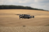A drone is flying over an expansive, golden field, likely a wheat field, with gentle rolling hills in the background. The sky appears cloudy, with soft lighting casting a serene glow over the landscape.