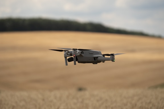 A drone is flying over an expansive, golden field, likely a wheat field, with gentle rolling hills in the background. The sky appears cloudy, with soft lighting casting a serene glow over the landscape.