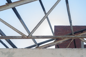 A partially constructed building with exposed metal beams forming a triangular pattern against a clear blue sky. Red bricks and a white concrete wall are visible, showing signs of weathering and rust on the metal framework.