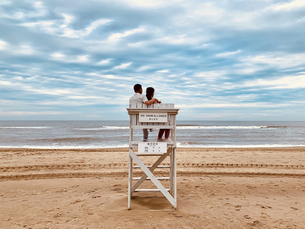 person sitting on white wooden lifeguard house on beach during daytime