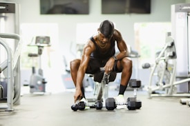 A person in athletic attire sits on a bench in a gym setting, reaching down to pick up a dumbbell from a row of dumbbells on the floor. Various gym equipment, such as treadmills and weight machines, are visible in the background.