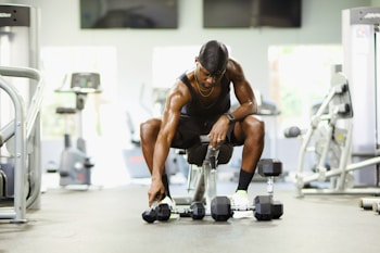 A person in athletic attire sits on a bench in a gym setting, reaching down to pick up a dumbbell from a row of dumbbells on the floor. Various gym equipment, such as treadmills and weight machines, are visible in the background.