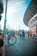 People are walking and cycling near a large stadium with a modern design. The scene includes a bicycle with orange accents leaning against a pole, and many people engaging in recreational activities. The sky is partly cloudy with tall city buildings visible in the background.