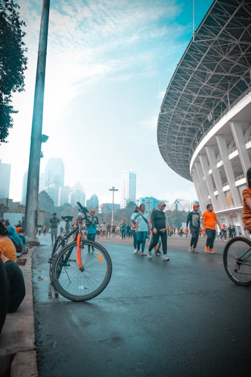 People are walking and cycling near a large stadium with a modern design. The scene includes a bicycle with orange accents leaning against a pole, and many people engaging in recreational activities. The sky is partly cloudy with tall city buildings visible in the background.