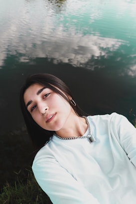 A person with dark hair and a nose piercing is wearing a white shirt and a chain necklace with a lock. They are lying on their back next to a calm body of water, with clouds reflected on the water's surface.