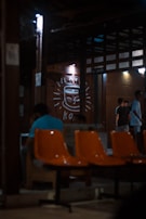 A dimly lit indoor setting with orange chairs in the foreground. There are people sitting and standing, engaged in conversations. The background features a large drawing of a smiling coffee cup on the wall, illuminated by small lights. The structure has a rustic and cozy appearance with wooden beams and walls.