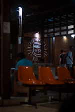 A dimly lit indoor setting with orange chairs in the foreground. There are people sitting and standing, engaged in conversations. The background features a large drawing of a smiling coffee cup on the wall, illuminated by small lights. The structure has a rustic and cozy appearance with wooden beams and walls.