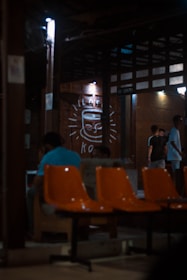 A dimly lit indoor setting with orange chairs in the foreground. There are people sitting and standing, engaged in conversations. The background features a large drawing of a smiling coffee cup on the wall, illuminated by small lights. The structure has a rustic and cozy appearance with wooden beams and walls.