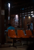 A dimly lit indoor setting with orange chairs in the foreground. There are people sitting and standing, engaged in conversations. The background features a large drawing of a smiling coffee cup on the wall, illuminated by small lights. The structure has a rustic and cozy appearance with wooden beams and walls.