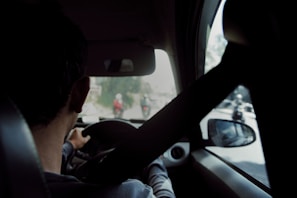A person is driving a car with a seatbelt fastened. The view is from the back seat, showing their hands on the steering wheel. Outside the front windshield, two motorcyclists are visible on a blurry road.