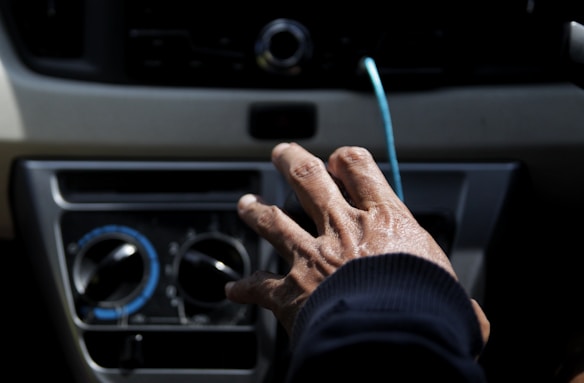 A close-up of a hand reaching towards the dashboard controls of a vehicle, with the hand appearing to adjust the settings on a panel. The background includes climate control knobs with marked temperature indications and surrounding dashboard elements.