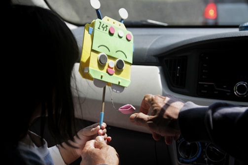 A person with a shadowed face holds a stick puppet with a green, cartoonish, vehicle-like character at the top. The character has a happy expression with large circular eyes and a smiling mouth. Another person's hand is interacting with the puppet, emphasizing a playful and interactive setting inside a car.