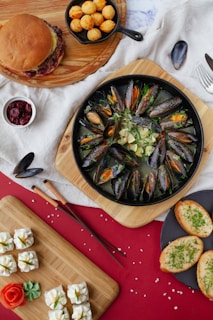 A diverse spread of food items is arranged on a table. There's a wooden platter with a hamburger and crispy fried balls next to a bowl of cranberries. A dish features mussels in a broth with slices of lemon and herbs, surrounded by slices of garlic bread. On another wooden board, small decorative desserts, resembling white roses, and an intricately crafted tomato rose are placed. Chopsticks rest beside the board, adding an elegant touch.