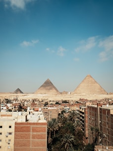 A cityscape with several buildings in the foreground, set against the backdrop of the Pyramids of Giza in the distance. The sky is mostly clear with a few scattered clouds, creating a contrasting view between urban architecture and ancient historical monuments.
