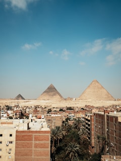 A cityscape with several buildings in the foreground, set against the backdrop of the Pyramids of Giza in the distance. The sky is mostly clear with a few scattered clouds, creating a contrasting view between urban architecture and ancient historical monuments.